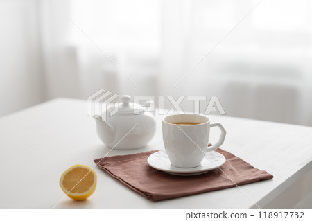 A cup of tea with lemon and a teapot on a white table against the background of a kitchen window. 118917732