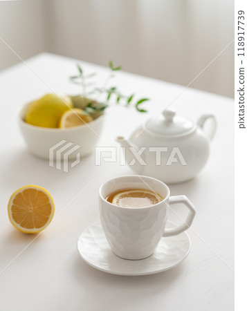 A cup of tea with lemon and a teapot on a white table against the background of a kitchen window. A cup of tea with lemon and a teapot on a white table against the background of a kitchen window. 118917739