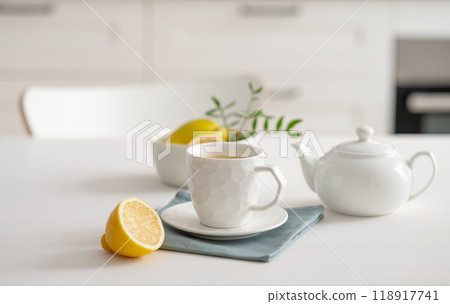 A cup of tea with lemon and a teapot on a white table against the background of a white kitchen. A cup of tea with lemon and a teapot on a white table against the background of a white kitchen. 118917741