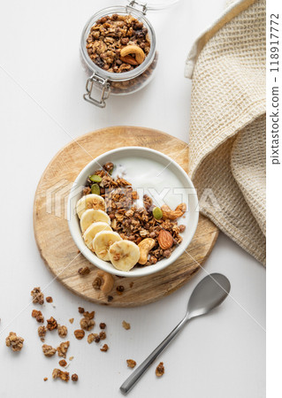 Granola with banana and nuts on a wooden board on a white table with a napkin and spoon. Granola with banana and nuts on a wooden board on a white table with a napkin and spoon. 118917772