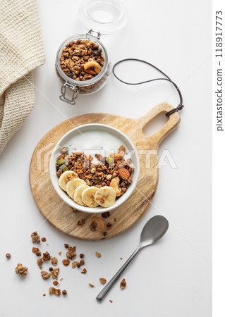 Granola with banana and nuts on a wooden board on a white table with a napkin and spoon. 118917773