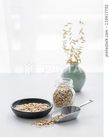 Oatmeal in a plate, scoop and in a jar on a white textured table in front of the kitchen window. Oatmeal in a plate, scoop and in a jar on a white textured table in front of the kitchen window. 118917782