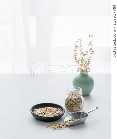 Oatmeal in a plate, scoop and in a jar on a white textured table in front of the kitchen window. Oatmeal in a plate, scoop and in a jar on a white textured table in front of the kitchen window. 118917784