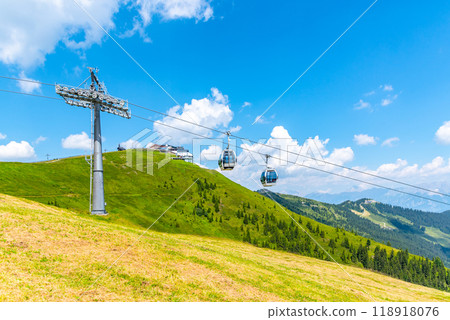 Visitors ride the Schmittenhohe cableway, enjoying stunning views of the Austrian Alps. The vibrant green landscape stretches below under a bright blue sky dotted with clouds. Visitors ride the Schmittenhohe cableway, enjoying stunning views of the Austrian Alps. The vibrant green landscape stretches below under a bright blue sky dotted with clouds. 118918076