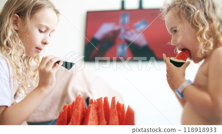 Two little sisters eat juicy red watermelon slices and watch TV together at home Two little sisters eat juicy red watermelon slices and watch TV together at home 118918933