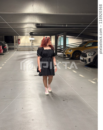 Millennial woman in a black dress walks through an empty parking garage, her pink sneakers adding a pop of color. Setting is urban and modern, with cars lined up neatly in the dimly lit space Millennial woman in a black dress walks through an empty parking garage, her pink sneakers adding a pop of color. Setting is urban and modern, with cars lined up neatly in the dimly lit space 118920368