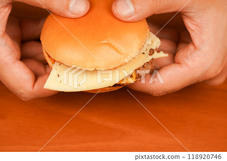 Home delivery. Man holds burger with hands. Hands holding homemade beef burger on wooden table. Ready to eat tasty fast food hamburger. Unhealthy food concept. Close up shot of young man eats burger. 118920746