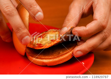 Close up shot of young man eats burger. Home delivery. Man holds burger with hands. Hands holding homemade beef burger on wooden table. Ready to eat tasty fast food hamburger. Unhealthy food concept. 118920787