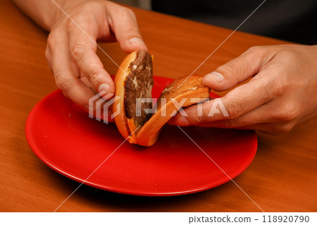 Man holds burger with hands. Hands holding homemade beef burger on wooden table. Ready to eat tasty fast food hamburger. Unhealthy food concept. Home delivery. Close up shot of young man eats burger. Man holds burger with hands. Hands holding homemade beef burger on wooden table. Ready to eat tasty fast food hamburger. Unhealthy food concept. Home delivery. Close up shot of young man eats burger. 118920790