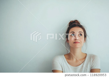 A young woman with a thoughtful expression sitting against a plain wall while contemplating her next decision 118920953