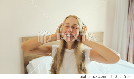 Happy smiling mature woman enjoying listening to music with headphones on the bed at home Happy smiling mature woman enjoying listening to music with headphones on the bed at home 118921504
