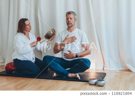 A man meditating with a sound bowl, and woman holds it during a calming sound therapy session A man meditating with a sound bowl, and woman holds it during a calming sound therapy session 118921804