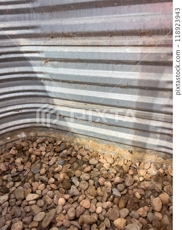 A detailed view of a corrugated metal window well with signs of rust and small holes. Gravel is scattered at the base, indicating outdoor exposure and wear over time.  118923943