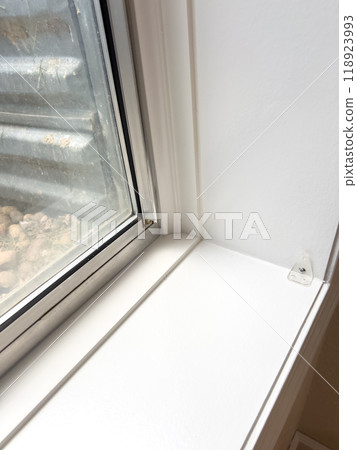 A close-up view of a basement window showing the white interior window frame, with an outside view of a metal window well lined with small rocks. The clean window trim and well-maintained frame 118923993