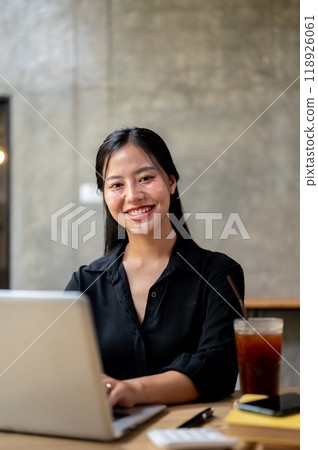 An attractive Asian woman working remotely from a coffee shop, sitting at a table with her laptop. An attractive Asian woman working remotely from a coffee shop, sitting at a table with her laptop. 118926061
