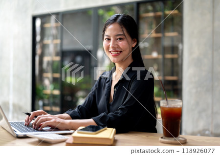 Attractive businesswoman sits at a desk indoors, smiling at the camera while working on her laptop. 118926077
