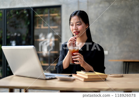 A beautiful Asian businesswoman enjoys iced coffee while working on her laptop, working remotely. A beautiful Asian businesswoman enjoys iced coffee while working on her laptop, working remotely. 118926078