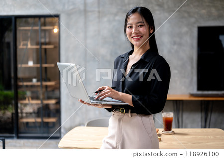 A confident Asian businesswoman is standing in her modern loft office with a laptop in her hand. 118926100