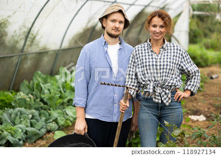 Man and woman with rakes in hands inside greenhouse with vegetable harvest Man and woman with rakes in hands inside greenhouse with vegetable harvest 118927134