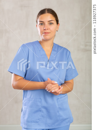 Friendly nurse in blue coat posing against gray studio background, looking at camera with smile Friendly nurse in blue coat posing against gray studio background, looking at camera with smile 118927375