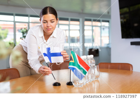 Positive young woman putting little flag of Finland on table next to the flag of South Africa Positive young woman putting little flag of Finland on table next to the flag of South Africa 118927397
