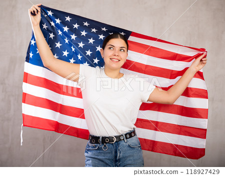 Joyful young woman waving national flag of USA flag while standing against gray wall indoors Joyful young woman waving national flag of USA flag while standing against gray wall indoors 118927429