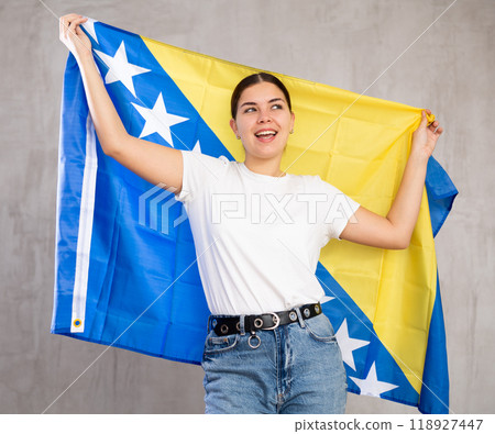 Happy young female with flag of Bosnia and Herzegovina against background of gray wall Happy young female with flag of Bosnia and Herzegovina against background of gray wall 118927447