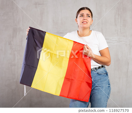 Stressed out sad young female holding flag of Belgium against gray wall background 118927570