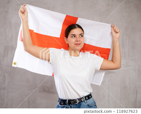Excited young woman in casual wear holding flag of England against gray wall background Excited young woman in casual wear holding flag of England against gray wall background 118927623