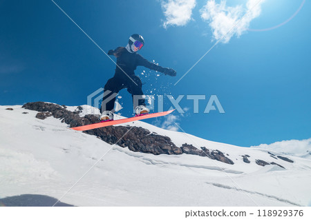 Female snowboarder jumping off a ski jump and doing a trick. Amazing shot of a snowboarder flying in the air and grabbing a trick against the backdrop of snowy mountains 118929376