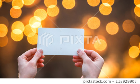 Close-up hand holding mockup white card ready for branding, against backdrop of soft glowing bokeh lights, business or personal graphic design 118929905