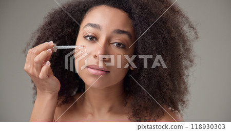 Beauty and healthcare concept - beautiful african american woman with curly afro hairstyle and clean, healthy skin applies skin care serum from a pipette to her skin Beauty and healthcare concept - beautiful african american woman with curly afro hairstyle and clean, healthy skin applies skin care serum from a pipette to her skin 118930303