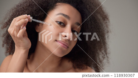 Beauty and healthcare concept - beautiful african american woman with curly afro hairstyle and clean, healthy skin applies skin care serum from a pipette to her skin Beauty and healthcare concept - beautiful african american woman with curly afro hairstyle and clean, healthy skin applies skin care serum from a pipette to her skin 118930307