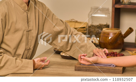A practitioner of traditional medicine checks a patient's veins by placing the patient's hand on a towel-covered wooden board, with required supplies and medication packets arranged around the table. 118932507