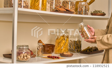 A pharmacist examines a glass jar that contains dried herbs, including licorice root, in this scene, which is set in a traditional Chinese medicine clinic with tidy shelves. A pharmacist examines a glass jar that contains dried herbs, including licorice root, in this scene, which is set in a traditional Chinese medicine clinic with tidy shelves. 118932521