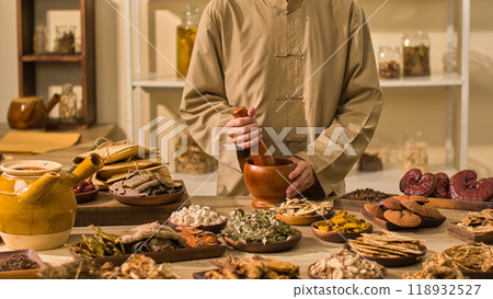 In a laboratory setting for a traditional medicine photo, a doctor dressed in brown grinds dry herbs with a wooden mortar and pestle while other herbal materials surround a table. 118932527