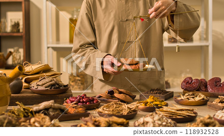 A medical practitioner weighs reishi mushrooms on a traditional scale, while other herbs and laboratory tools are arranged around the table, template picture for traditional medicine advertisements. A medical practitioner weighs reishi mushrooms on a traditional scale, while other herbs and laboratory tools are arranged around the table, template picture for traditional medicine advertisements. 118932528