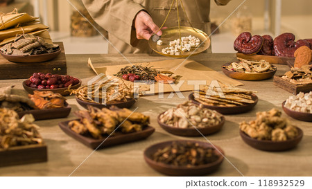 The conventional health care photo template depicts a scientist feeding Angelica sinensis blocks into brown paper wrappers containing other herbs, surrounded by other natural ingredients on a table. 118932529