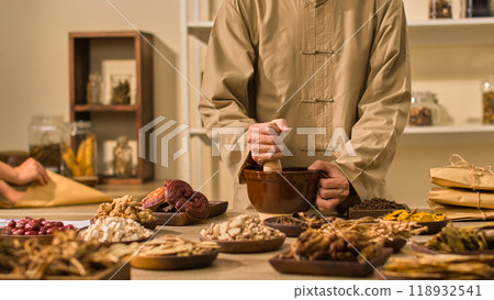 Two herbalists are pounding herbs in a mortar and taking notes beside it. On the table are herbal substances on wooden plates. Photo backdrop in traditional medical room for herbal content. 118932541