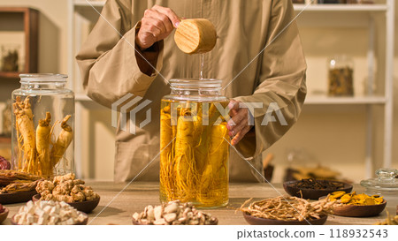 A photo sample with a background of a herbal chamber gets shown. A herbalist pours water from a ginseng jar and sets it on a table next to some dried plants. A photo sample with a background of a herbal chamber gets shown. A herbalist pours water from a ginseng jar and sets it on a table next to some dried plants. 118932543