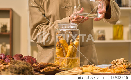 In a traditional medicine clinic, a practitioner pours water into a ginseng bottle as other herbal components are distributed on wooden trays. High quality photo template 118932546
