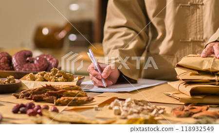 The herbalist is taking notes and inspecting the wrapped packages of herbal medicine. Image sample is situated a table covered in a variety of plants is located in the herbal medicine chamber. 118932549