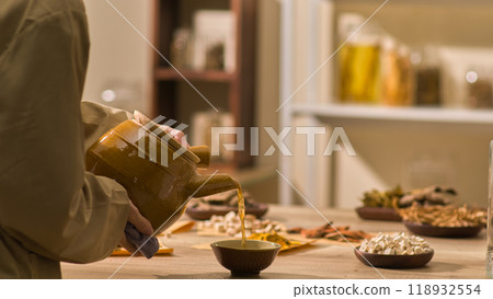 A traditional beige-suited herbalist is filling a ceramic cup with herbal water from a closet as other herbs are arranged on wooden platters. A charming oriental medical room with a template picture 118932554