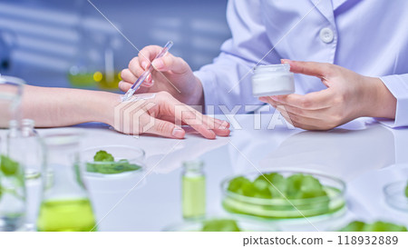 A scientist holding a white cream applies it to a person's arm, demonstrating the effectiveness of the cream. The background is a bright and clean laboratory with pennywort and scientific equipment. 118932889