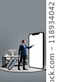 Businessman with suitcase pointing to large 3D model of phone with blank screen, representing seamless business travel and mobile convenience for professionals on the go. 118934042