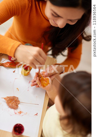 Cute little girl and mom spending good time painting hands and autumn yellow leaves together Cute little girl and mom spending good time painting hands and autumn yellow leaves together 118936564