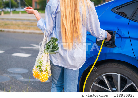 Blonde woman with long hair connecting an electric car to the charger, with a bag of groceries in her other hand, back view. Blonde woman with long hair connecting an electric car to the charger, with a bag of groceries in her other hand, back view. 118936923