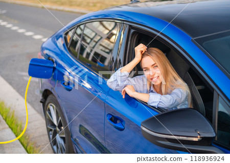 A woman enjoys her time in the passenger seat of an electric vehicle, charging in a serene outdoor setting. 118936924