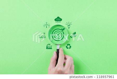 Green Energy and Sustainability Concept. Hand holding a magnifying glass focusing on a target with a leaf, surrounded by green energy and sustainability icons on a green background. Carbon footprint, 118937961