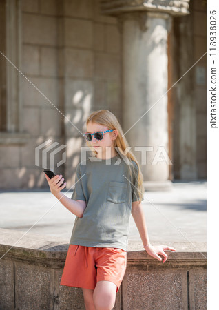 Young girl in sunglasses and with blond hair with a smartphone in her hands stands on the street Young girl in sunglasses and with blond hair with a smartphone in her hands stands on the street 118938026